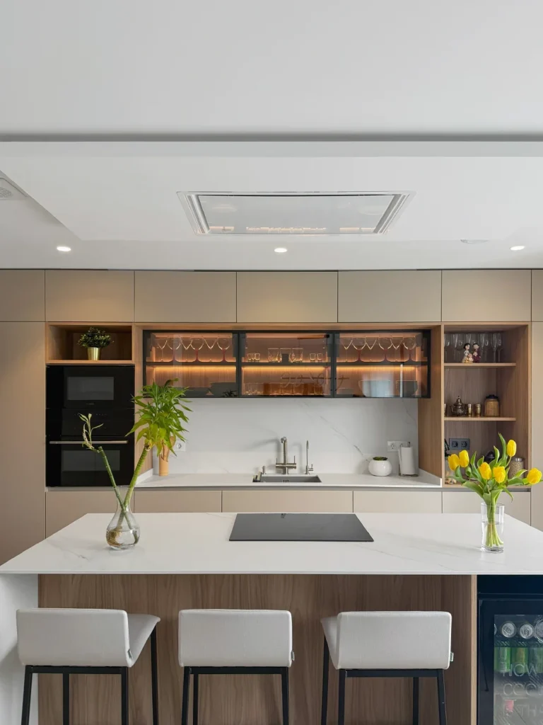Vista frontal de cocina con isla central, taburetes, mobiliario en tonos neutros y vitrinas iluminadas.
